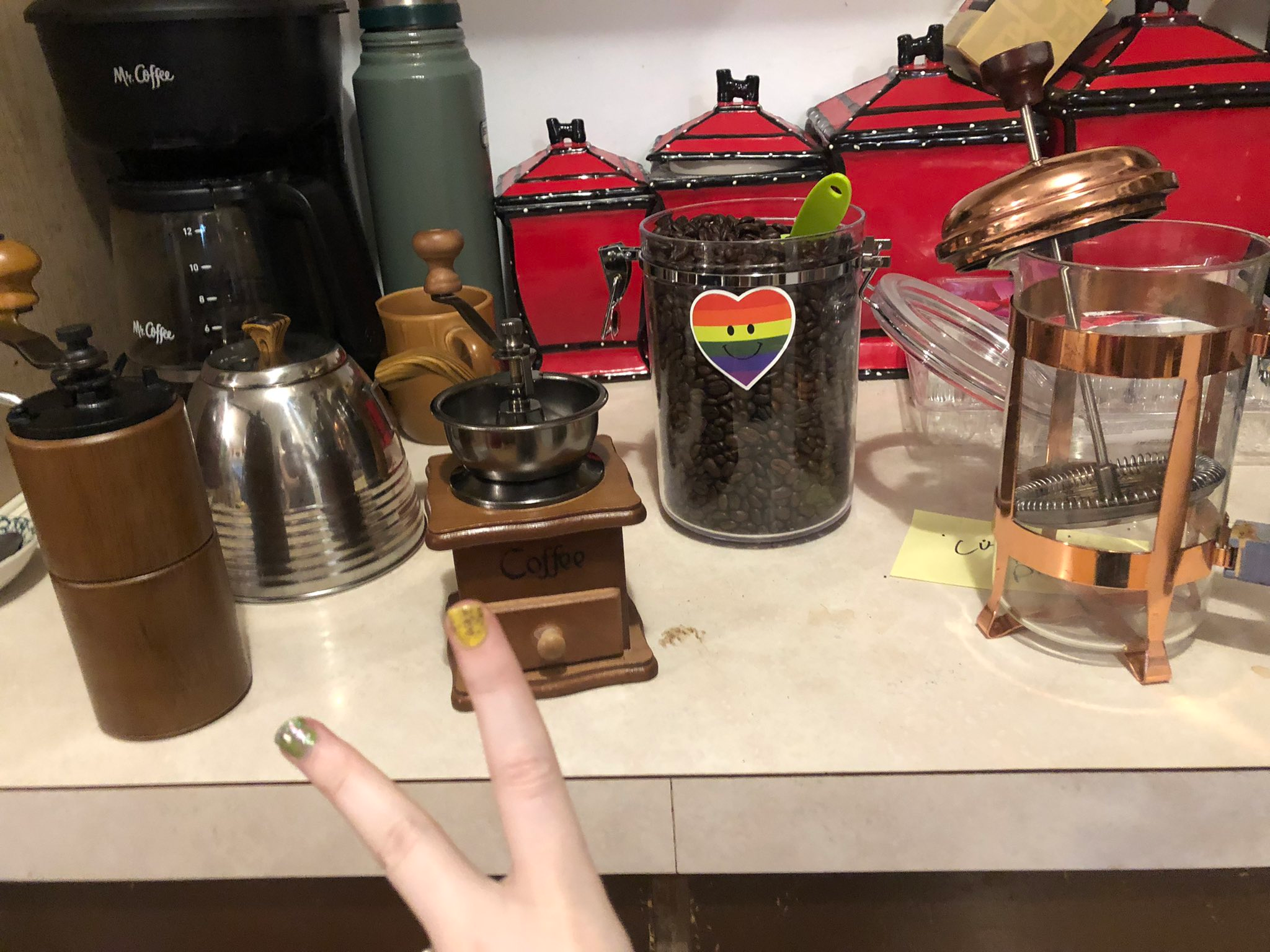 a copper french press, a wooden burr handheld grinder, a steel gooseneck kettle, a small tabletop wooden burr grinder, a clear snap canister of coffee beans with gay pride stickers, and a kitchen scale barely in frame.